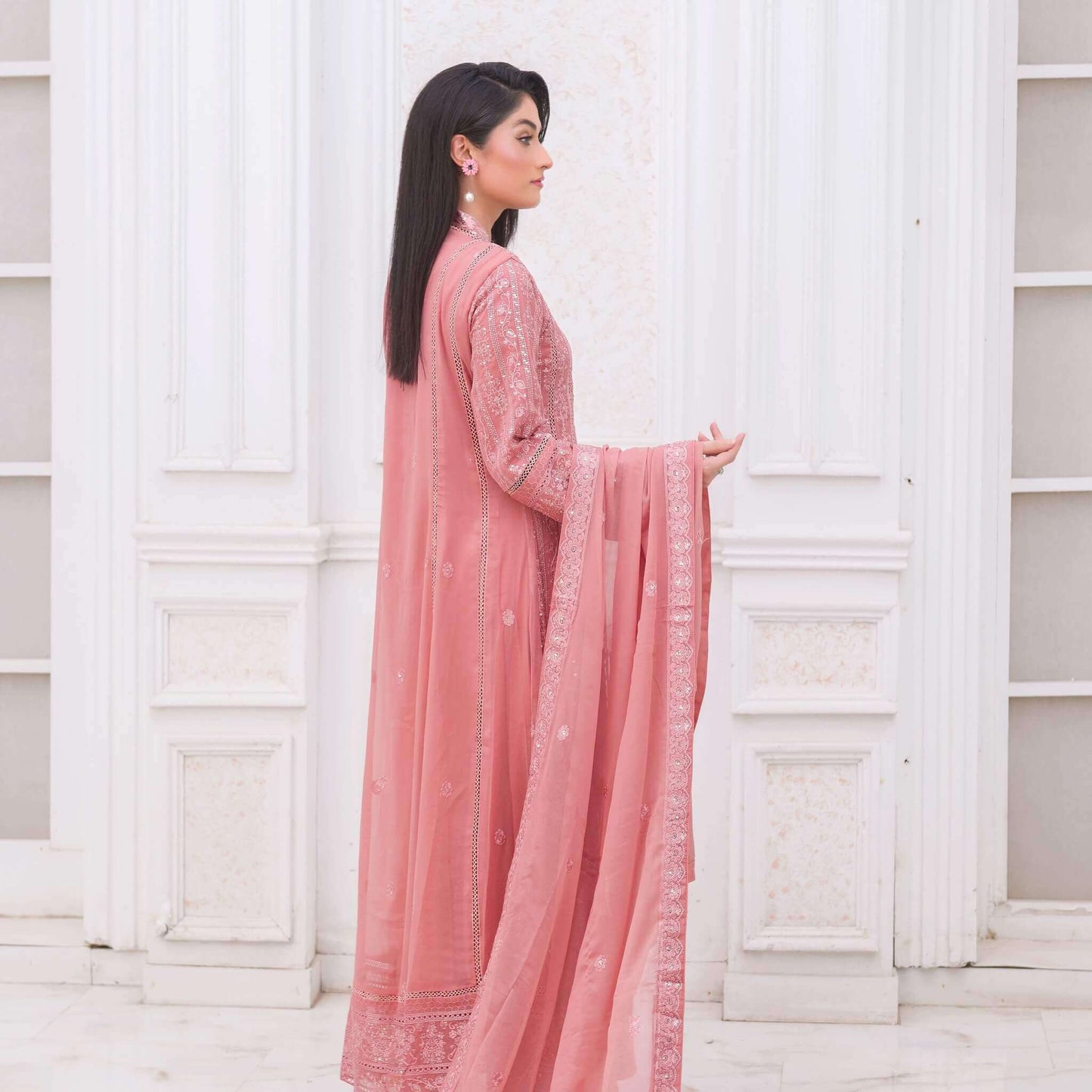 Woman in a pink traditional outfit standing against a white paneled wall back pose