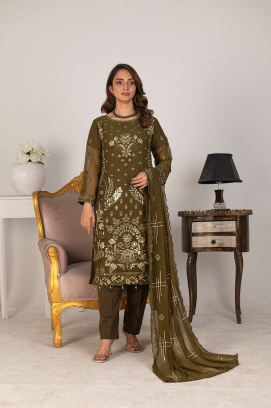 Woman in a green embroidered outfit standing in a room with a chair and table.